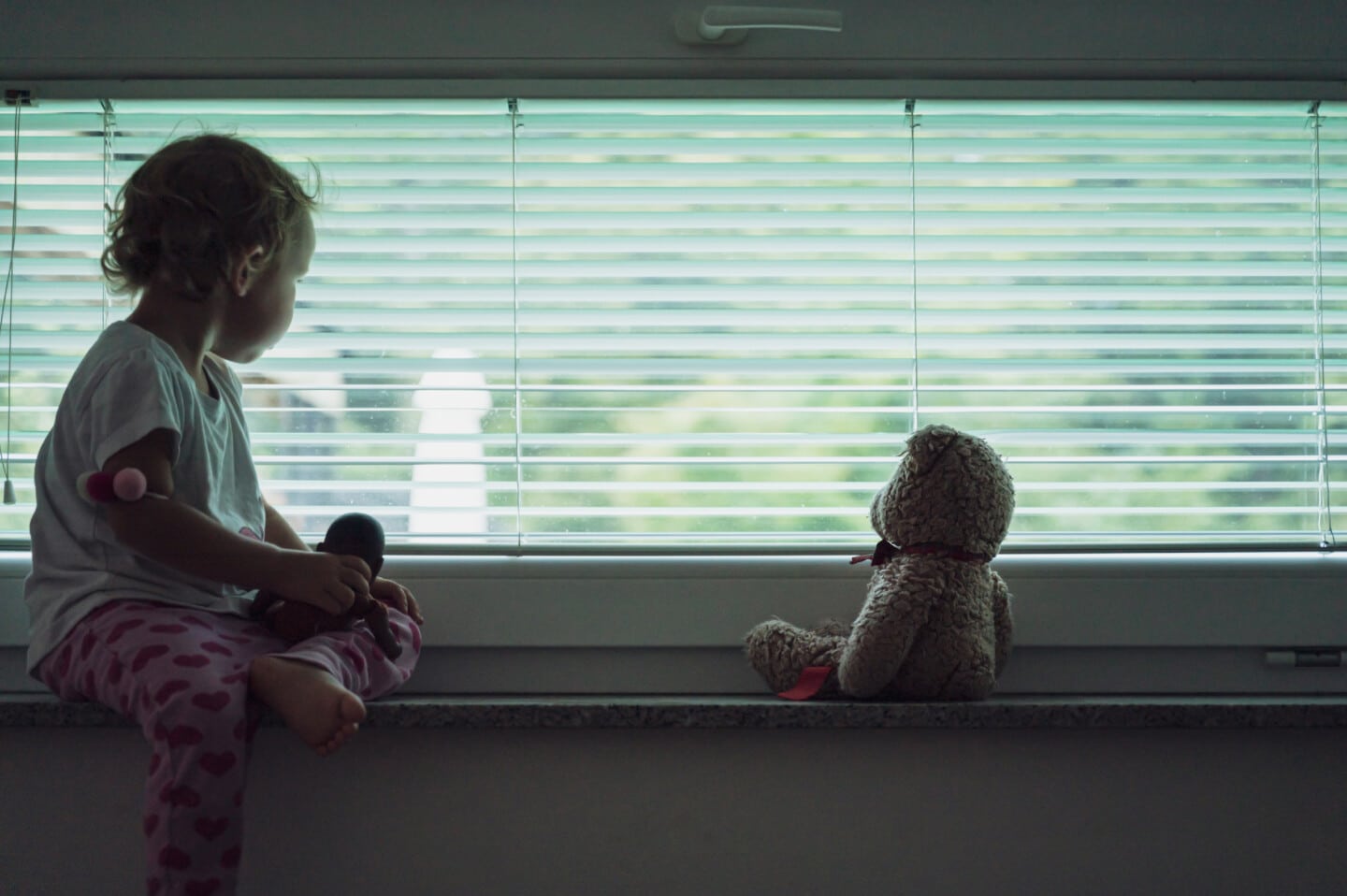 Toddler girl sitting on window shelf  with her teddy bear looking out the window with blinds on it. Conceptual image of child abuse and abandonment.