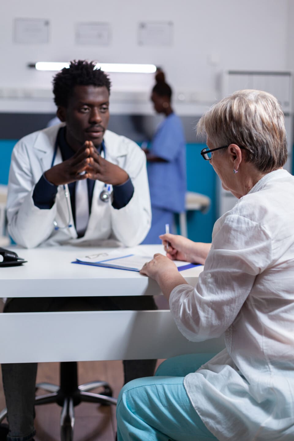 Doctor of african ethnicity consulting elderly patient sitting at white desk in office workplace. Black man with uniform and old woman discussing healthcare treatment and medicine